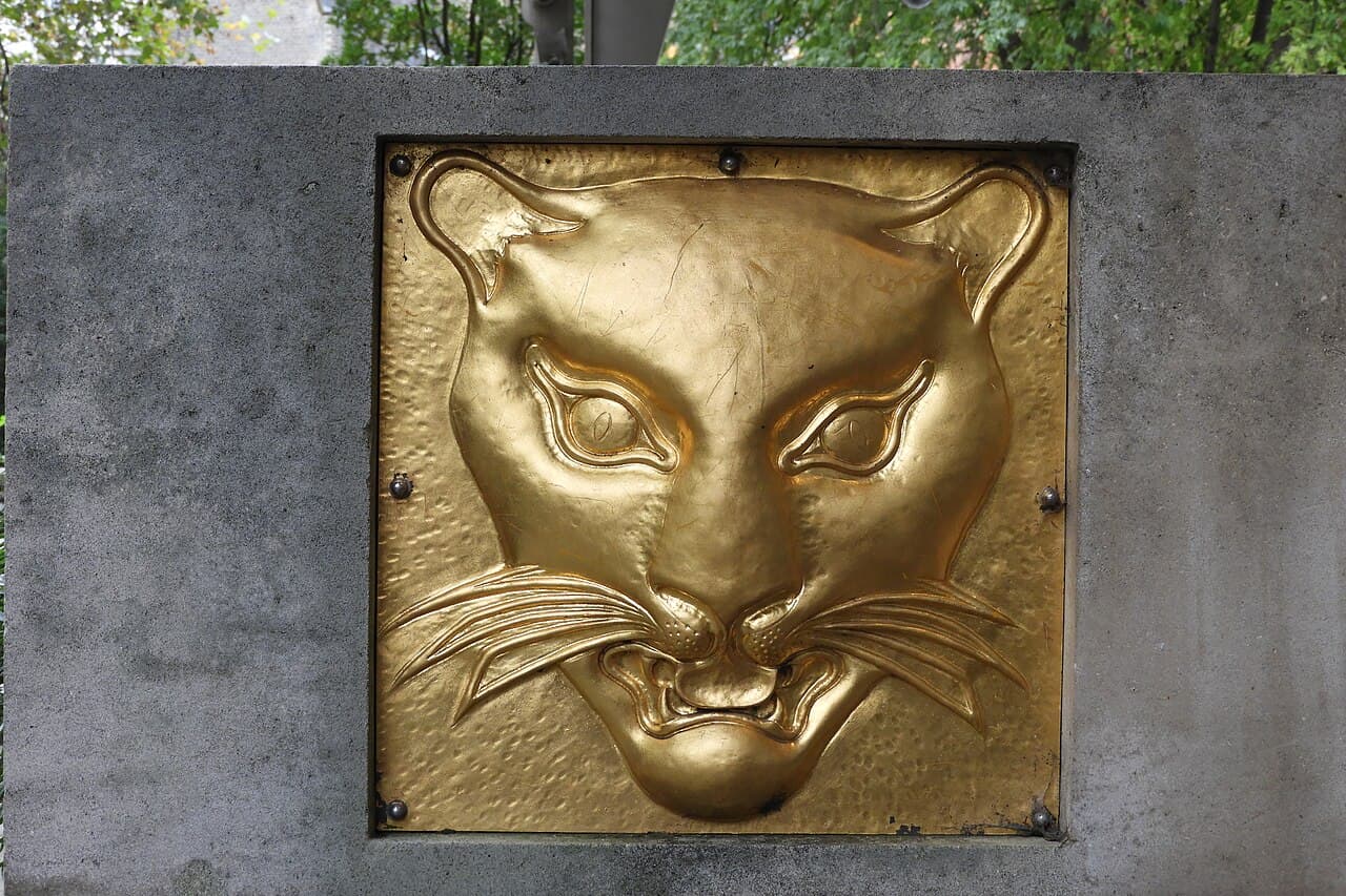 A gilded relief of a leopard's face set into a grey stone block, with trees behind.